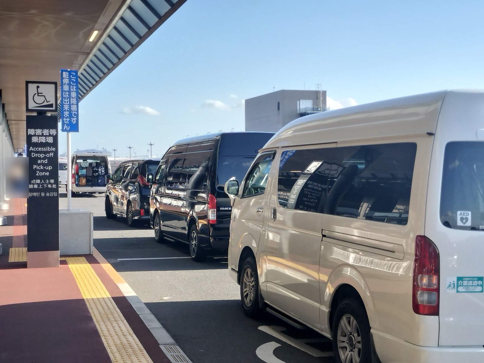 成田空港の障害者等乗降場（Accessible Drop-off and Pick-up Zone）の案内看板と、乗降場に並ぶ福祉車両